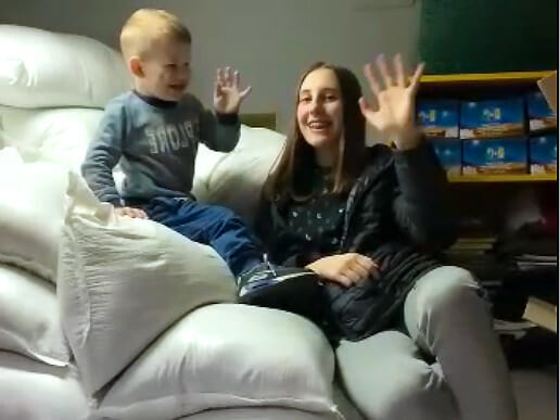 A woman and a child are sitting on a bed and waving.