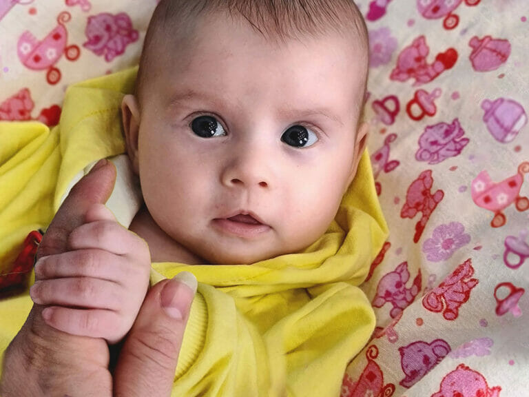 A baby in a yellow blanket holding a hand on a Mission Possible.