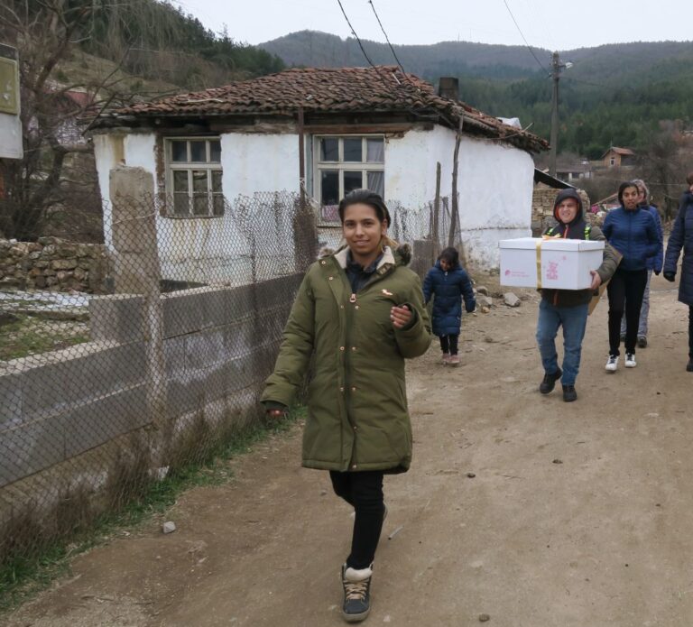 A group of people walking along a dirt road, one carrying a large white box, in front of a small house with a tiled roof.