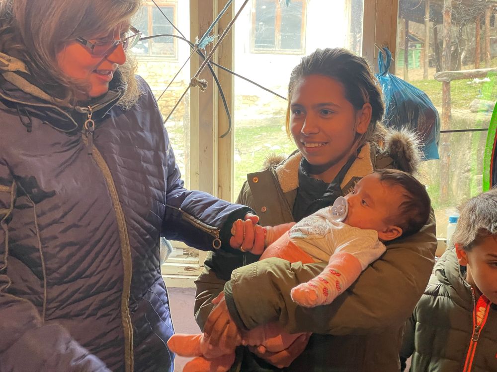 An older woman reaching to hold the hand of an infant being held by a young woman.